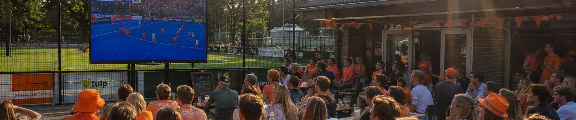 Supporters kijken tijdens een zonnige middag samen naar een WK Hockey-wedstrijd op een groot professioneel scherm op het terras van een hockeyclub.